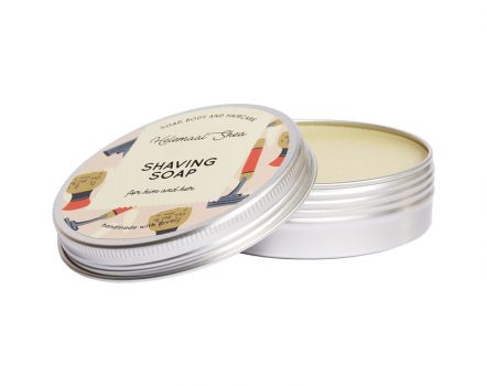 A tin of HelemaalShea's Shaving Soap with the lid partially open, revealing the soap inside. The label features the product name and a decorative design.