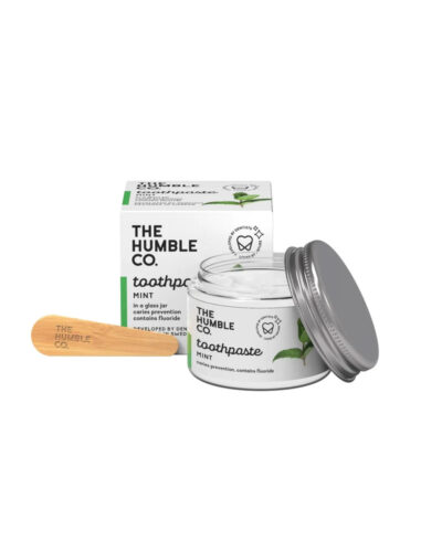 The Humble Co mint Toothpaste. Glass jar with aluminum lid open to the side, next to compostable wooden spoon. Recyclable paper packaging the in the background