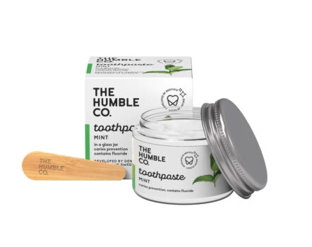 The Humble Co mint Toothpaste. Glass jar with aluminum lid open to the side, next to compostable wooden spoon. Recyclable paper packaging the in the background