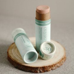 A plastic-free tinted sunstick SPF 50 by Odyskin, displayed with its eco-friendly, compostable packaging. The sunstick has a light brown tint and is shown standing upright, with its lid off, next to an open box and a storage bag. The packaging features a minimalist design in pastel green with white text, emphasizing its natural and eco-friendly attributes.